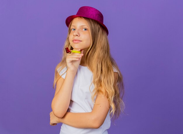 Bambina graziosa in cappello di vacanza che tiene il fischio che guarda l'obbiettivo sorridente con la faccia felice, concetto di festa di compleanno in piedi su sfondo viola