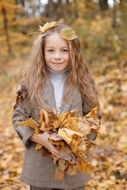 Bambina in abiti di moda che cammina nel parco d'autunno. Ragazza che tiene foglie gialle. Ragazza che indossa un costume marrone con giacca.