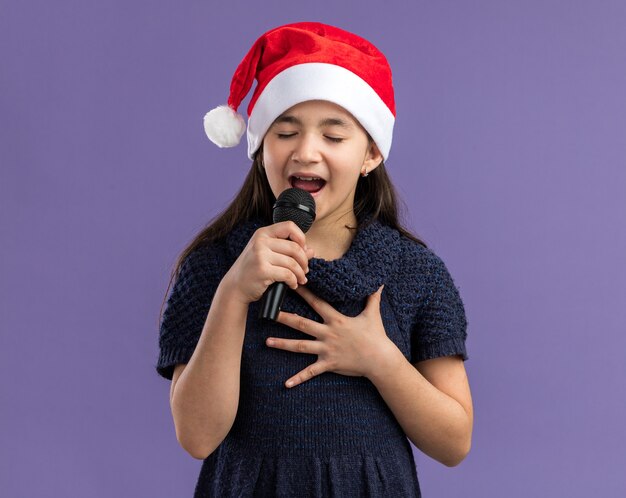 Bambina in abito a maglia che indossa cappello da Babbo Natale che tiene il microfono che canta celebrando la festa di Natale felice e positiva in piedi sul muro viola