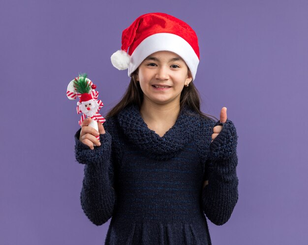 Bambina in abito a maglia che indossa un cappello da Babbo Natale con in mano un bastoncino di zucchero natalizio che sorride allegramente in piedi sul muro viola