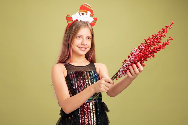 Bambina in abito da festa glitterato e fascia con Babbo Natale che tiene rami con bacche rosse che guardano da parte sorridendo allegramente in piedi su sfondo verde