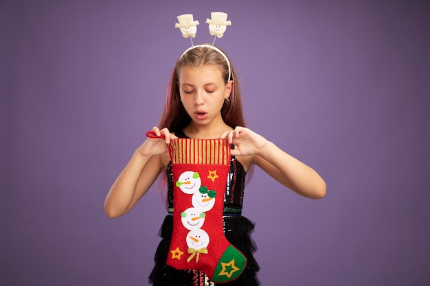 Bambina in abito da festa glitterato e fascia divertente che tiene la calza di Natale guardando dentro incuriosita in piedi su sfondo viola