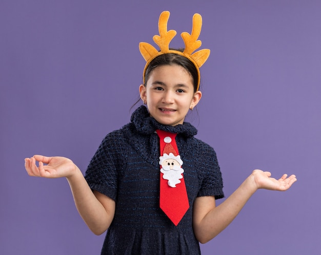 Bambina in abito di maglia che indossa cravatta rossa con bordo divertente con corna di cervo sulla testa sorridente confuso allargando le braccia ai lati in piedi sul muro viola