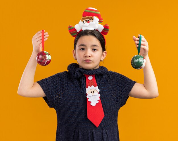 bambina in abito di maglia che indossa cravatta rossa con bordo divertente sulla testa tenendo le palle di Natale guardando la fotocamera con il viso serio in piedi su sfondo arancione