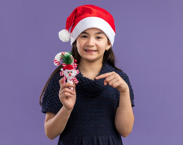 Bambina in abito in maglia che indossa cappello da Babbo Natale che tiene un bastoncino di zucchero natalizio che punta con il dito indice su di esso felice e positivo sorridente allegramente in piedi sul muro viola