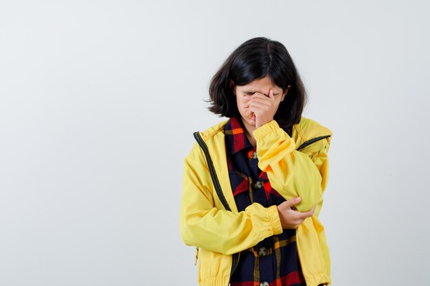 Bambina in camicia a quadri, giacca che copre il viso con la mano e sembra malinconica, vista frontale.