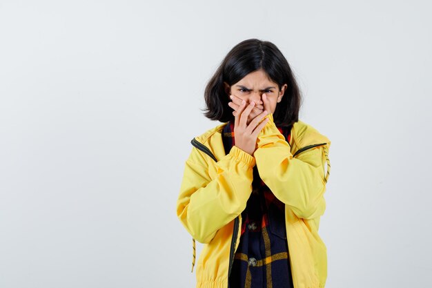 Bambina in camicia a quadri, giacca che tiene le mani sulla bocca e sembra stupita, vista frontale.