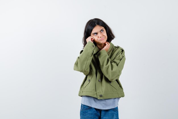 Bambina in cappotto, t-shirt, jeans che guarda lontano mentre pensa e sembra pensierosa, vista frontale.