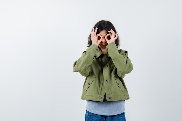 Bambina in cappotto, t-shirt, jeans che mostra il gesto degli occhiali e sembra carina, vista frontale.