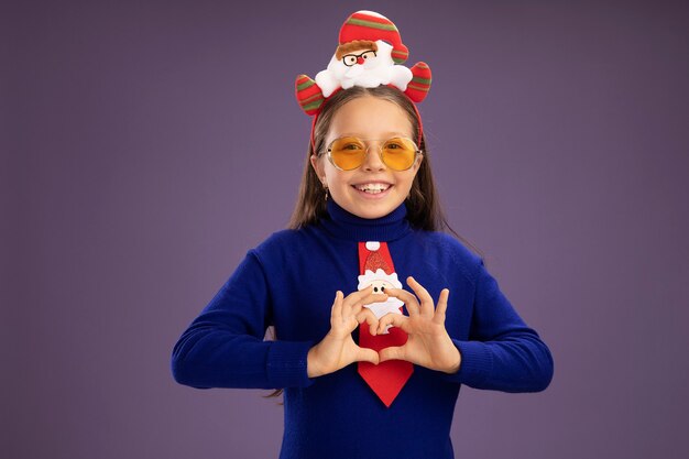 Bambina in dolcevita blu con cravatta rossa e divertente bordo di Natale sulla testa guardando la telecamera sorridendo allegramente facendo il gesto del cuore con le dita in piedi su sfondo viola