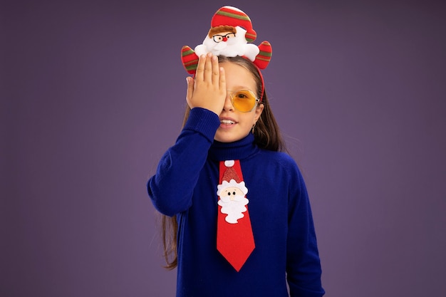 Bambina in dolcevita blu con cravatta rossa e divertente bordo natalizio sulla testa che copre un occhio con un braccio in piedi sul muro viola