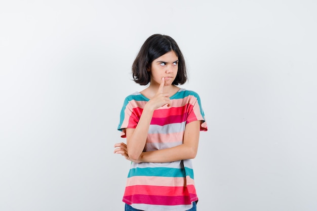 Bambina in maglietta che mette il dito indice vicino alla bocca, pensando a qualcosa e guardando pensieroso, vista frontale.