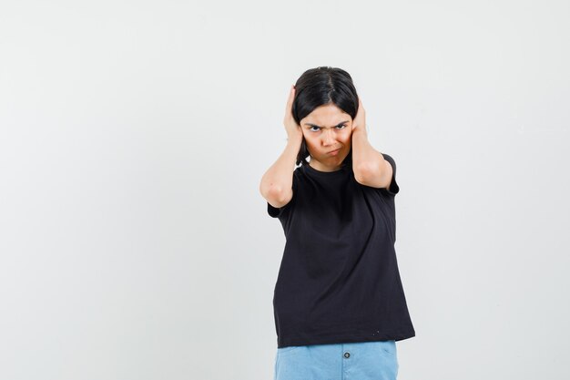 Bambina in maglietta nera, pantaloncini tenendo le mani sulle orecchie e guardando testardo, vista frontale.