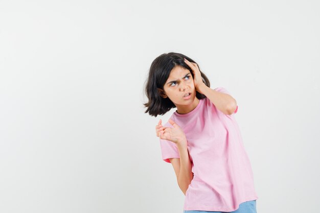 Bambina in maglietta rosa, pantaloncini tenendo la mano sulla testa e guardando offeso, vista frontale.