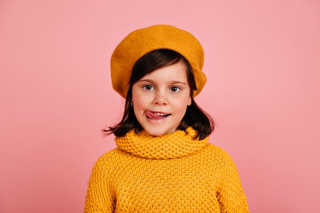 Bambina in maglione lavorato a maglia in piedi sulla parete rosa. bambino in posa con la lingua fuori.