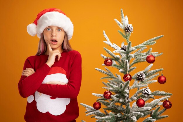 Bambina in maglione natalizio e cappello da Babbo Natale che guarda la telecamera stupita e sorpresa in piedi accanto a un albero di Natale su sfondo arancione