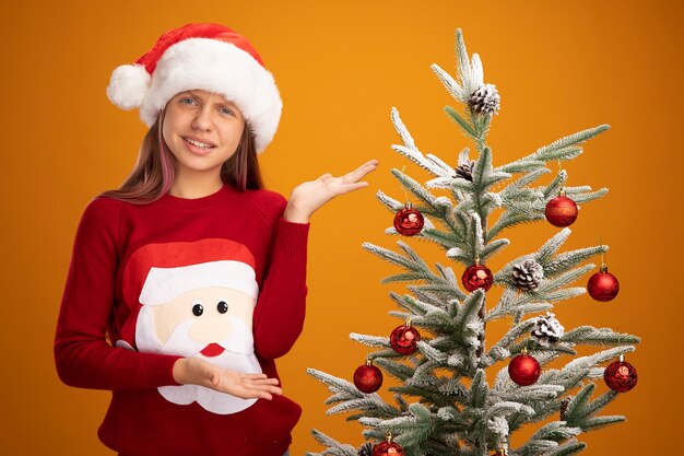 Bambina in maglione natalizio e cappello da Babbo Natale in piedi accanto a un albero di Natale che lo presenta con le braccia oh mani sorridenti confuse su sfondo arancione