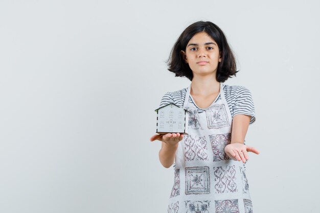 Bambina in t-shirt, grembiule che presenta il modello di casa e sembra sicura,