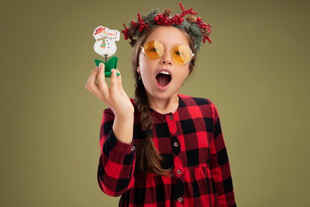 Bambina indossa la corona di Natale in abito controllato tenendo il giocattolo di Natale guardando la fotocamera in piedi felice ed eccitato su sfondo verde