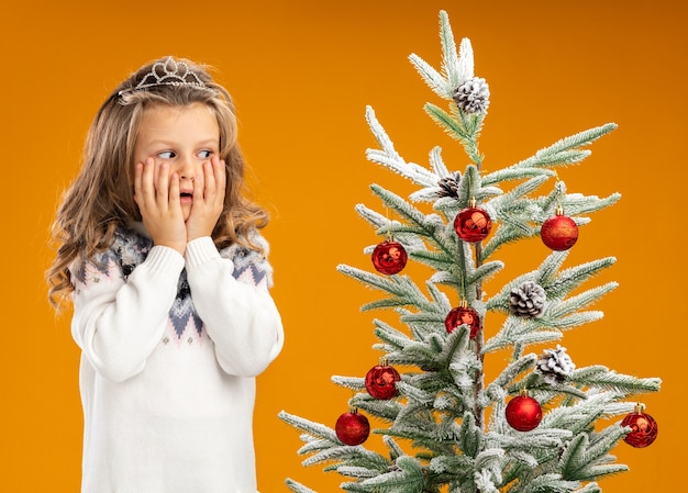 Bambina interessata che sta vicino all'albero di Natale che indossa la tiara con la ghirlanda sul collo ha coperto le guance con le mani isolate sulla parete arancione