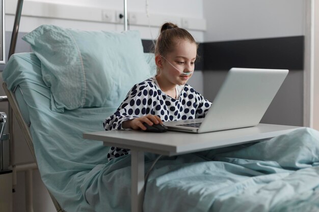 Bambina malata che gioca sul computer portatile mentre è ricoverata in ospedale nella stanza del reparto pediatrico della clinica sanitaria. Bambino malato che riposa sul letto del paziente mentre si diverte a giocare su un computer moderno.
