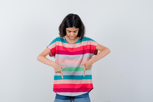 Bambina rivolta verso il basso con l'indice in t-shirt, jeans e guardando concentrata, vista frontale.