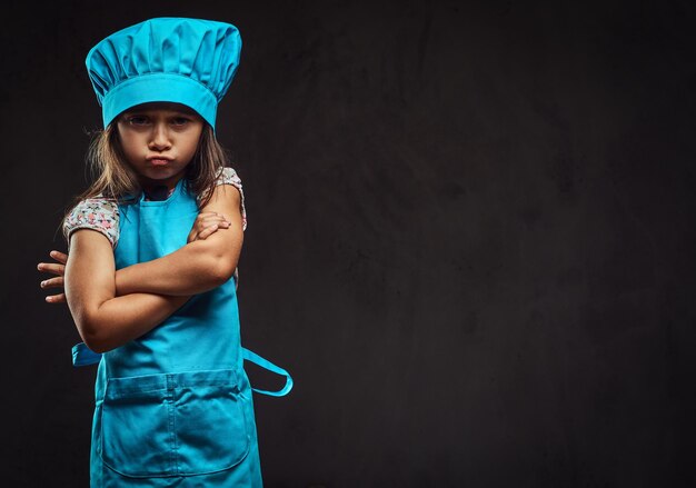 Bambina scontenta vestita in uniforme da cuoco blu in piedi con le braccia incrociate. Isolato su uno sfondo scuro con texture.