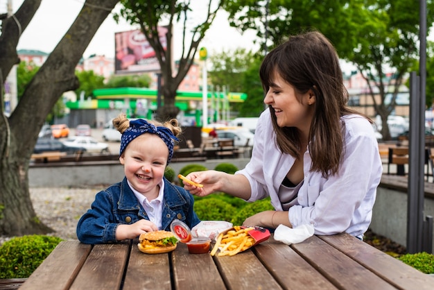 Bambina sveglia allegra che si siede con sua madre con hamburger e patatine fritte sul tavolo in un caffè