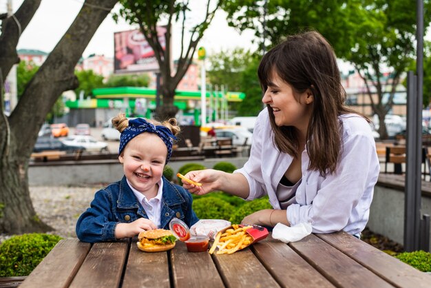 Bambina sveglia allegra che si siede con sua madre con hamburger e patatine fritte sul tavolo in un caffè