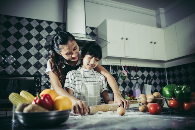 Bambina sveglia e sua madre in grembiuli che appiattiscono la pasta facendo uso di un matterello nella cucina