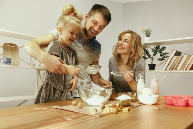 Bambina sveglia ed i suoi bei genitori che preparano la pasta per la torta in cucina a casa.