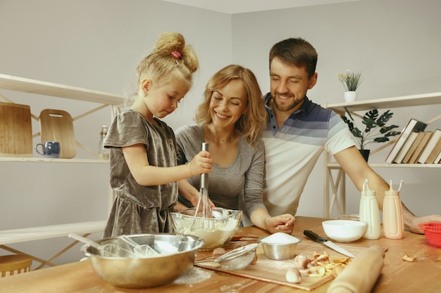 Bambina sveglia ed i suoi bei genitori che preparano la pasta per la torta in cucina a casa.