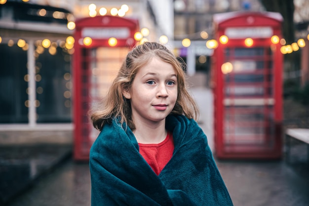 Bambina sveglia su uno sfondo sfocato di cabine telefoniche rosse e con bokeh