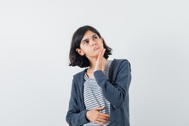 Bambina tenendo la mano sul mento in t-shirt, giacca e guardando pensieroso