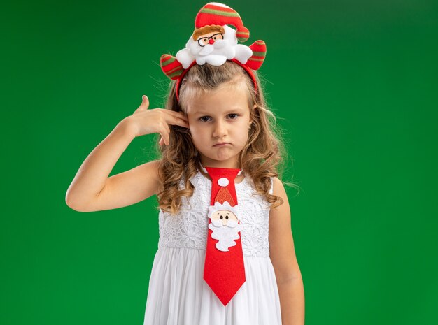 Bambina triste che indossa un cerchio per capelli di Natale con cravatta che graffia l'orecchio isolato sul muro verde