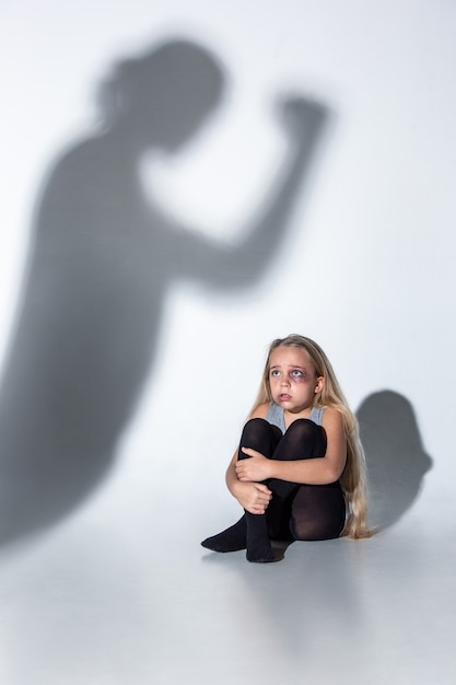 Bambina triste e spaventata con occhi iniettati di sangue e lividi che piange spaventata dall'ombra sul muro.