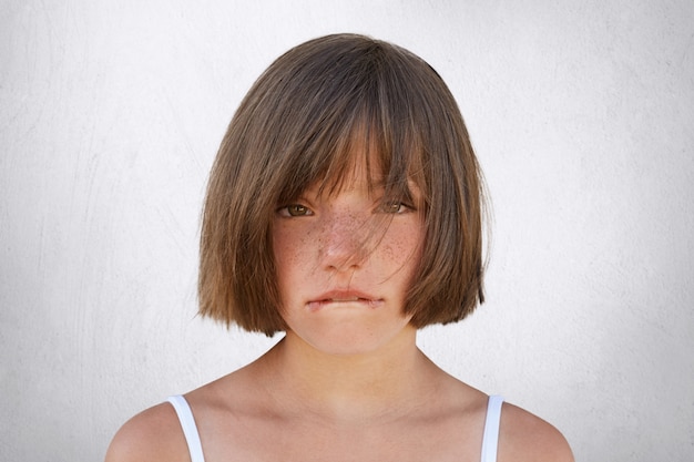 Bambina turbata che ha litigio con i suoi genitori, esaminante innocentemente nella macchina fotografica mentre curvando le sue labbra isolate su bianco. Bambina sorridente che piange, mordendosi il labbro inferiore