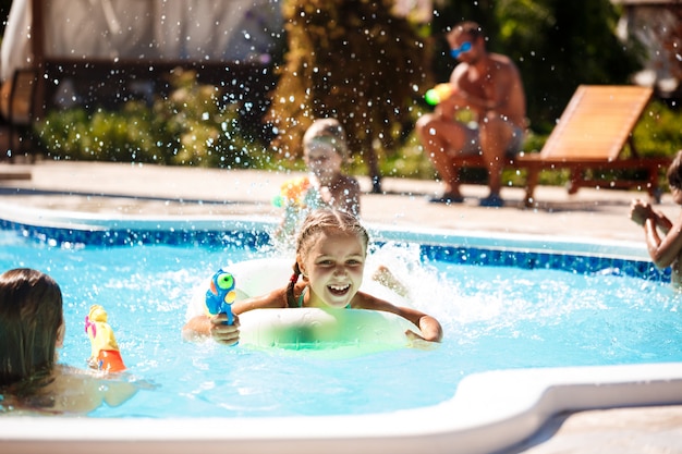 Bambini allegri che giocano a pistole ad acqua, gioendo, saltando, nuotando in piscina.