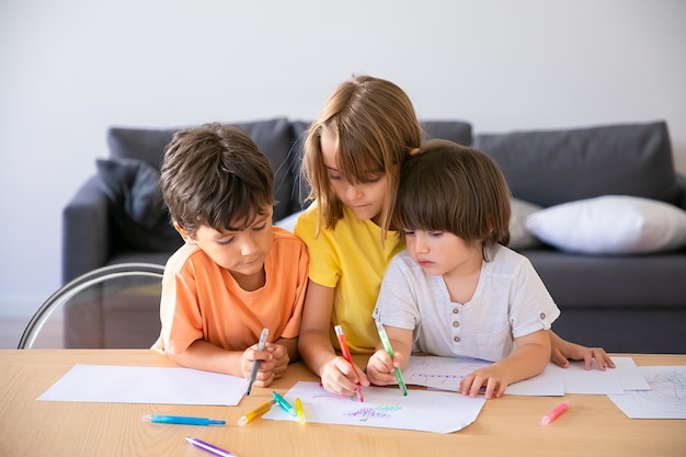 Bambini caucasici dipinto con pennarelli in soggiorno. Ragazzini svegli e ragazza bionda seduti a tavola insieme, disegno su carta e giocando in casa. Infanzia, creatività e concetto di fine settimana