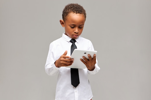 Bambini e concetto di tecnologia moderna. Scolaro afroamericano concentrato serio in uniforme che tiene compressa digitale generica bianca, gioco in linea o apprendimento, con espressione concentrata