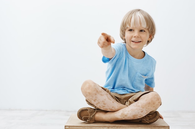 bambino biondo spensierato felice che viene intrattenuto, seduto con i piedi incrociati e indicando di lato mentre sorride ampiamente, essendo interessato e curioso