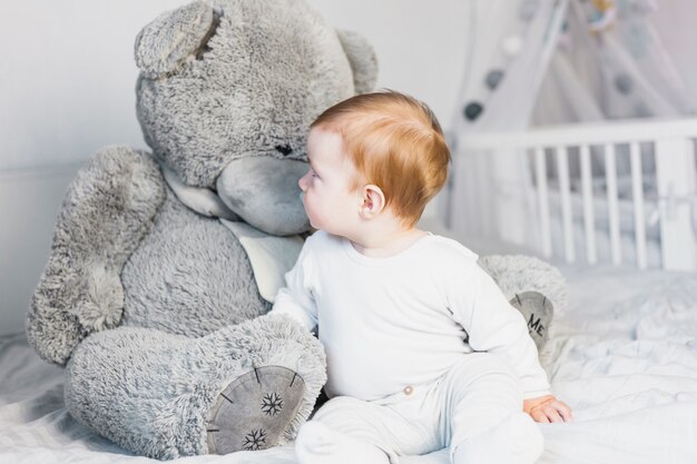 Bambino biondo sveglio in letto bianco con orsacchiotto