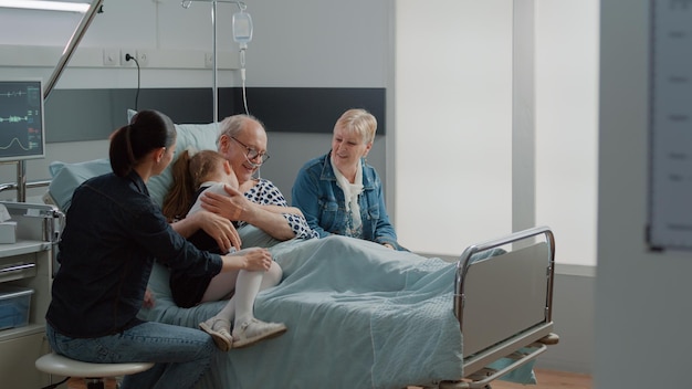 Bambino che abbraccia il nonno malato nel letto del reparto ospedaliero durante la visita, confortando il paziente anziano con malattia. Bambino e madre che visitano l'uomo anziano con la sacca antigoccia IV e il tubo dell'ossigeno nasale. Visitatori in clinica