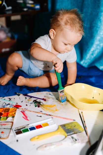 Bambino con la pittura della spazzola su carta vicino ai colori di acqua e sulla seduta sul coverlet