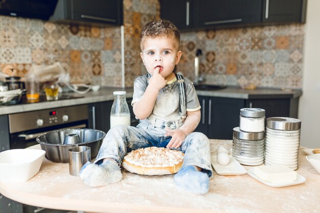 Bambino divertente che si siede sul tavolo della cucina in una cucina rustica che gioca con la farina e assaggia una torta.