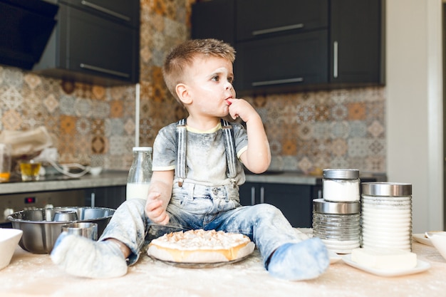 Bambino divertente che si siede sul tavolo della cucina in una cucina rustica che gioca con la farina e assaggia una torta.