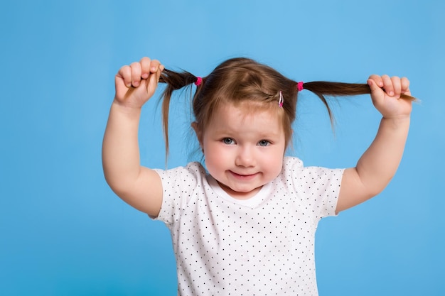 Bambino divertente in maglietta bianca su sfondo blu. Bambina graziosa che si aggrappa alle code. Copia spazio per il testo. Vendita, concetto di festa di compleanno.