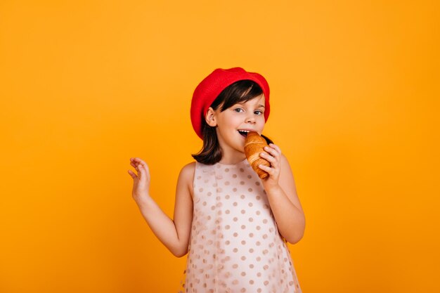 Bambino felice che mangia croissant con piacere Studio di una ragazza francese in posa su sfondo giallo