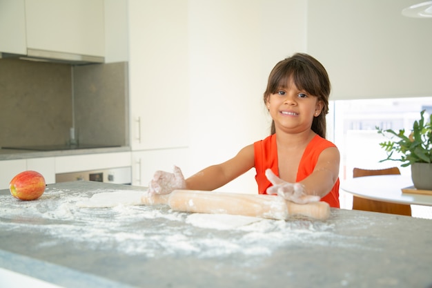 Bambino felice che rotola la pasta al tavolo della cucina da sola. Ragazza con farina sulle braccia che cuociono il pane o la torta. Colpo medio. Concetto di cucina familiare