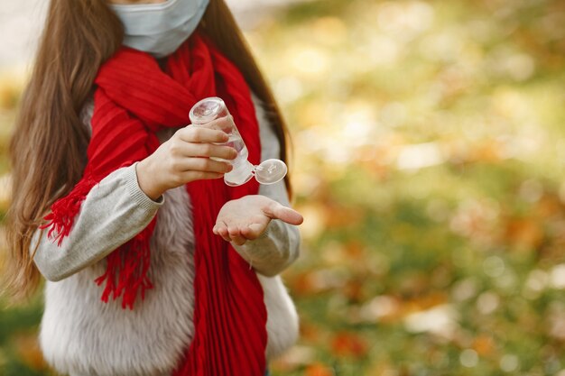 Bambino in piedi nella sosta di autunno. Tema Coronavirus. Ragazza in una sciarpa rossa. Il bambino usa l'antisettico.